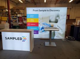 VK-1357 Backlit Exhibit with SEG Tension Fabric Graphics, MOD-1716 Backlit Custom Counter with Wireless Charging Pad and Locking Storage, and MOD-1454 Bistro Table with Wireless Charging Pads and LED Accent Lights